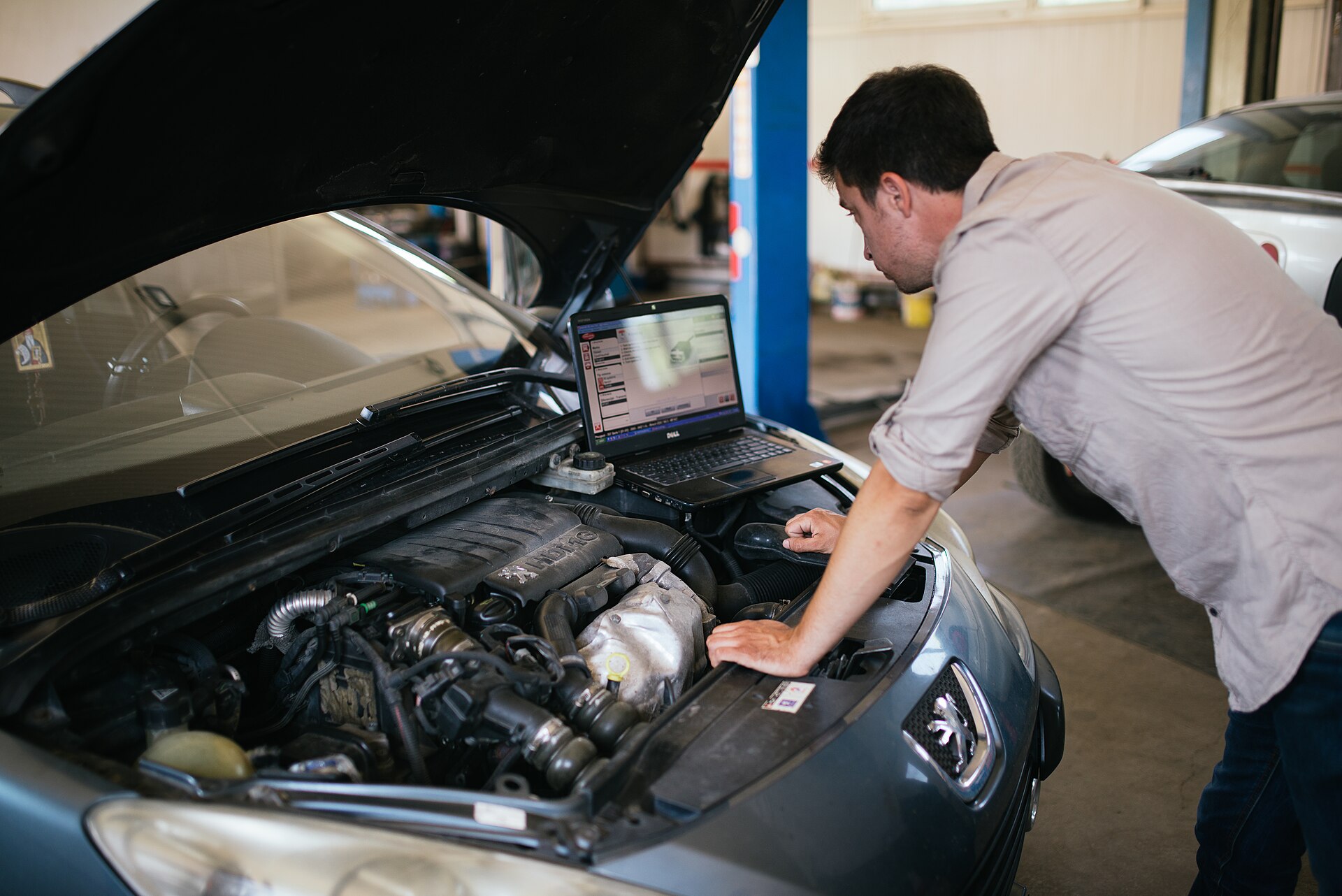 Techniker mit Laptop auf dem Kotflügel arbeitet am geöffneten Motorraum eines Fahrzeugs